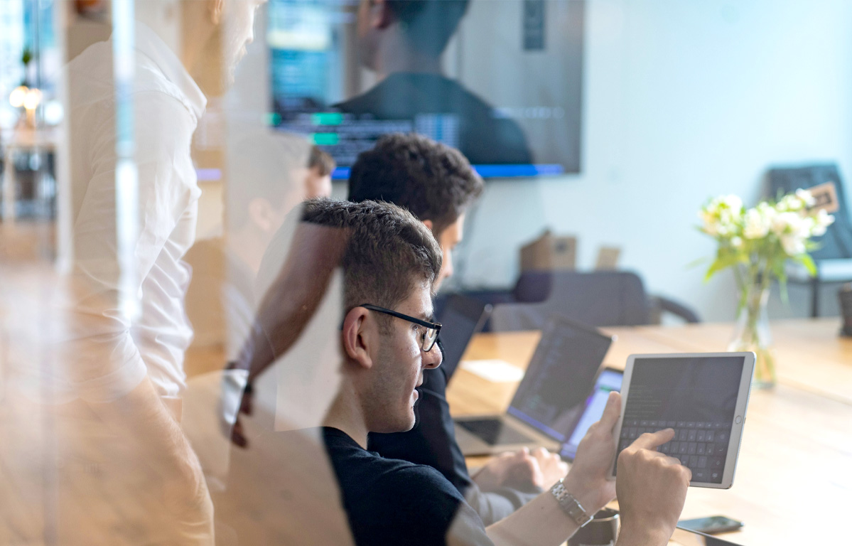 Drei Männer sitzen gemeinsam an einem Tisch und arbeiten konzentriert an ihren Computern. Hinter ihnen steht ein weiterer Mann in einem weißen Hemd, der über ihre Schultern blickt und ihre Arbeit überprüft.